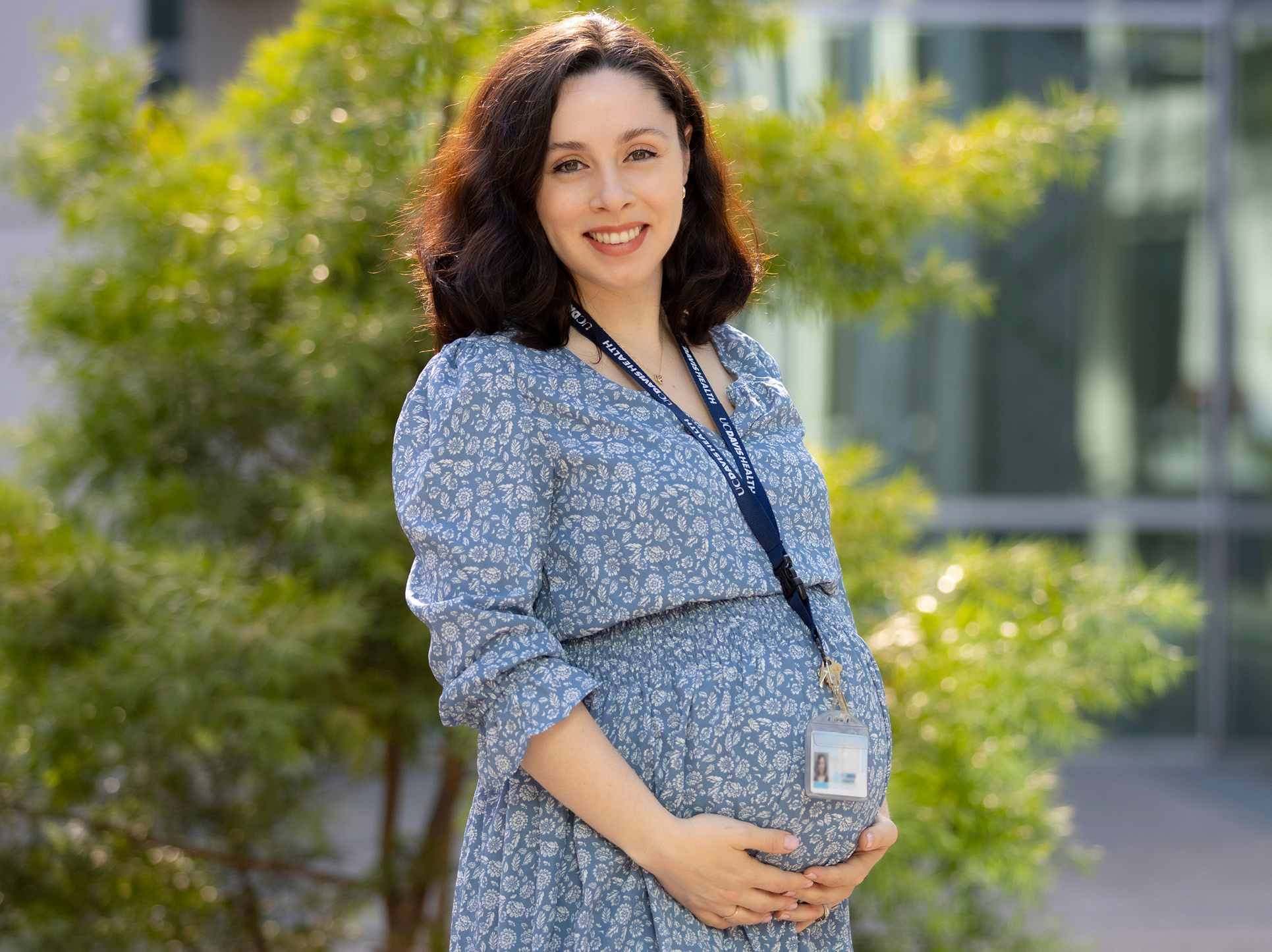 Photo of Caroline wearing a blue dress and a UC Davis Health badge, smiling at camera with hands loosely clasped at their stomach. 