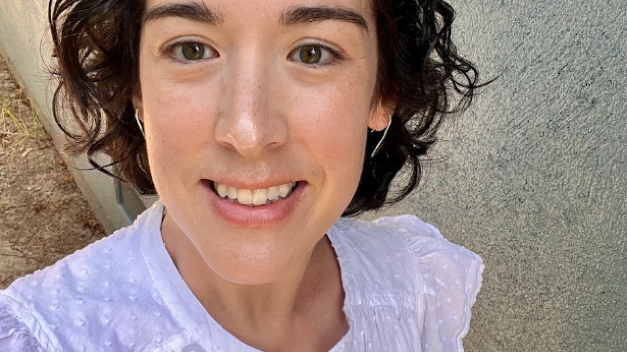 Smiling woman with curly hair wearing a white blouse, standing against a textured wall.