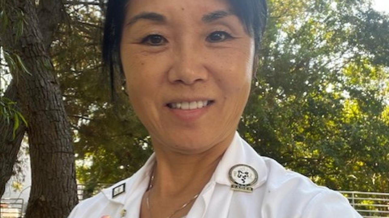 A healthcare professional in a white lab coat, smiling outdoors with greenery in the background.